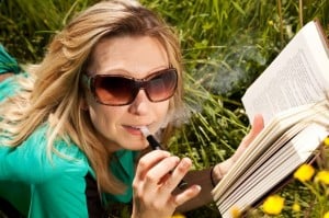 Blond kvinde med solbriller læser bog og ryger elektronisk cigaret
